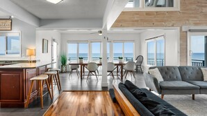 A spacious living area with a wooden floor, a grey sofa, and a dining table with chairs. - Pool, 7 Bed 7 Bath, Direct Oceanfront, 4 Kitchens (Port Orange)