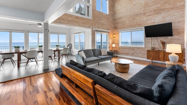 A spacious living area with a large sofa, a dining table with chairs, and a TV. The room has wooden walls and large windows offering a view of the sea. - Pool, 7 Bed 7 Bath, Direct Oceanfront, 4 Kitchens (Port Orange)