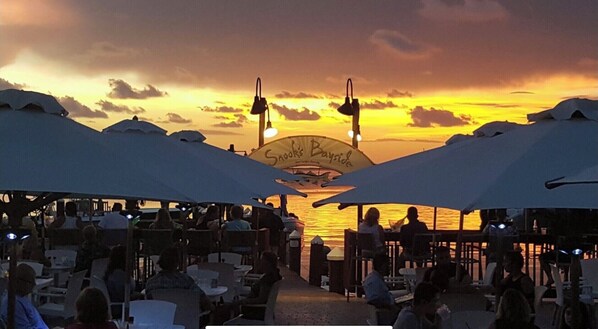 An outdoor dining area with umbrellas and tables, overlooking a body of water at sunset.
