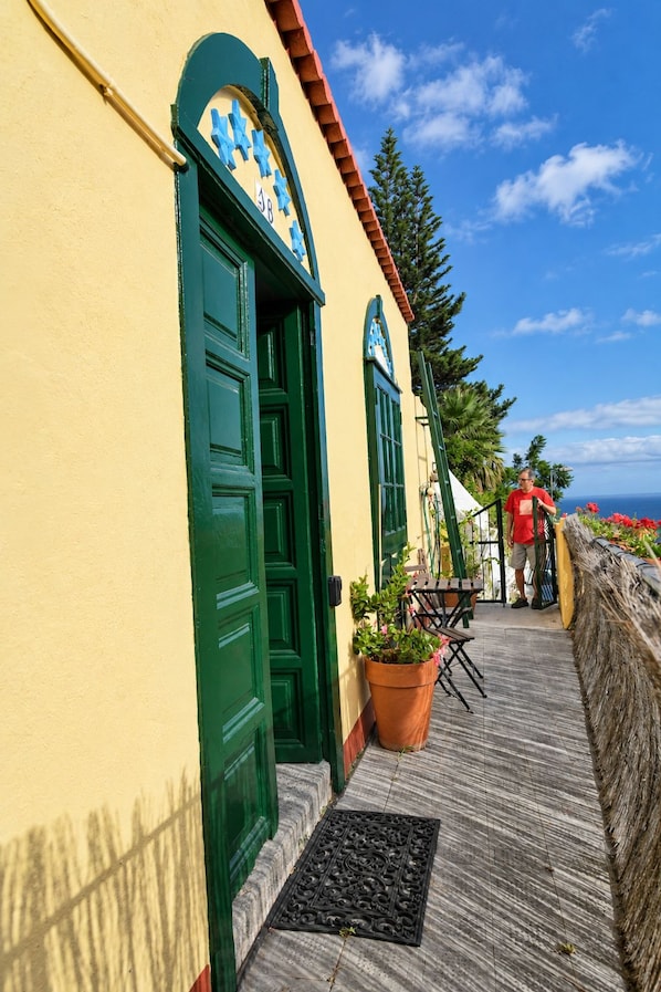 A building with green doors and windows, a tiled roof, and a small outdoor seating area with chairs and a table.