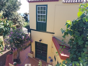 Un bâtiment à deux étages avec une fenêtre et une porte vertes, un petit balcon et un jardin agrémenté de fleurs violettes.