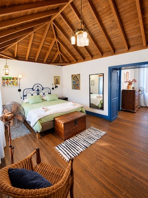 A bedroom with a wooden ceiling, a bed with green bedding, a bedside table, a standing fan, a wicker chair, and a large mirror.