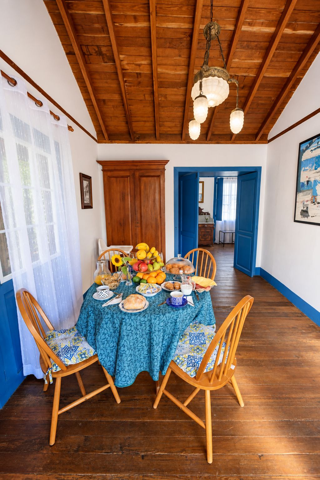 Een eetkamer met een houten plafond, een tafel gedekt met een blauwe tafelkleed en stoelen met gestreepte kussens.