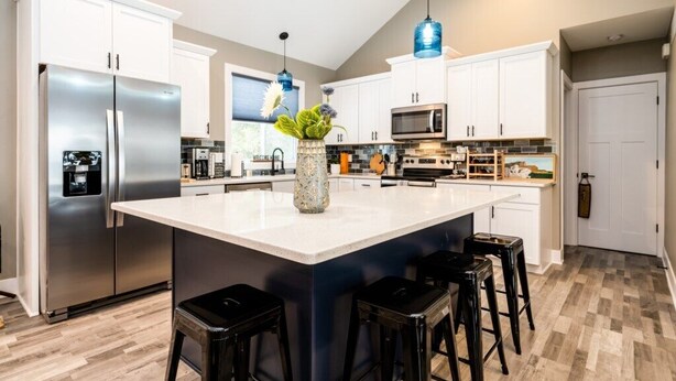 Una cocina moderna con armarios blancos, electrodomésticos de acero inoxidable, una isla central con taburetes y luces colgantes azules.