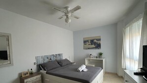 Una camera da letto con un letto, comodino, abat-jour, ventilatore a soffitto, un quadro, televisione e finestra con tende.