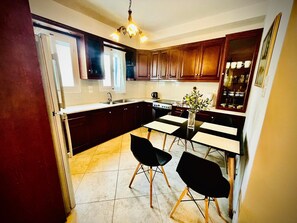 A compact kitchen with dark wooden cabinets, a central island, and a dining area with two black chairs. - Villa Arietta Santorini (Santorini)
