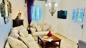 A living room with a sectional sofa, a wooden coffee table, a red vase, and a television on the wall. - Villa Arietta Santorini (Santorini)