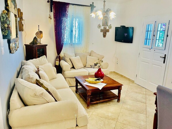 A living room with a sectional sofa, a wooden coffee table, a red vase, and a television on the wall. - Villa Arietta Santorini (Santorini)