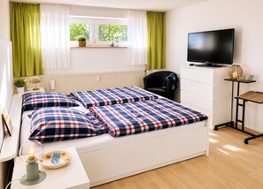 Ein Schlafzimmer mit Bett, Schreibtisch, Stuhl, Fernseher und einem Fenster mit Vorhängen.