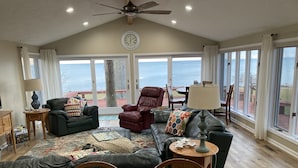 A spacious living room with a large window overlooking a scenic view, a ceiling fan, and a dining area with wooden furniture. - Spacious Lakefront Home with Amazing View (Huron)
