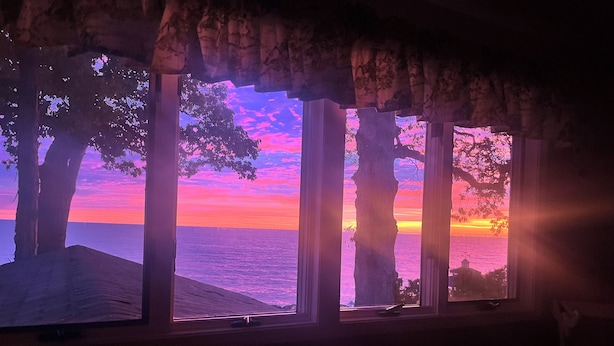 A room with a large window overlooking a beach at sunset. - Spacious Lakefront Home with Amazing View (Huron)