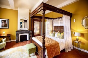 A four-poster bed with a canopy, a fireplace, a green armchair, and a small table with a lamp.