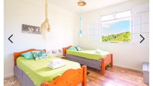 Une chambre avec deux lits en bois, une literie verte, un luminaire suspendu, une fenêtre donnant sur un paysage et un tableau au mur.