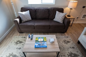 A brown sofa with leopard print cushions, a coffee table with a book and remote controls, and a lamp on a side table.