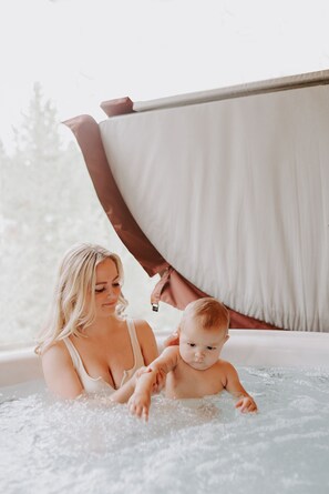 Une femme et un bébé dans un jacuzzi, avec vue sur les arbres à l’extérieur.