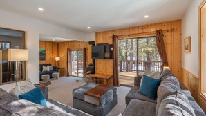 A modern living room with a sofa set, a coffee table, and a TV. There is a wooden deck outside with a view of trees. - Bernese Abode - Fireplace - Newly Updated! (Truckee)