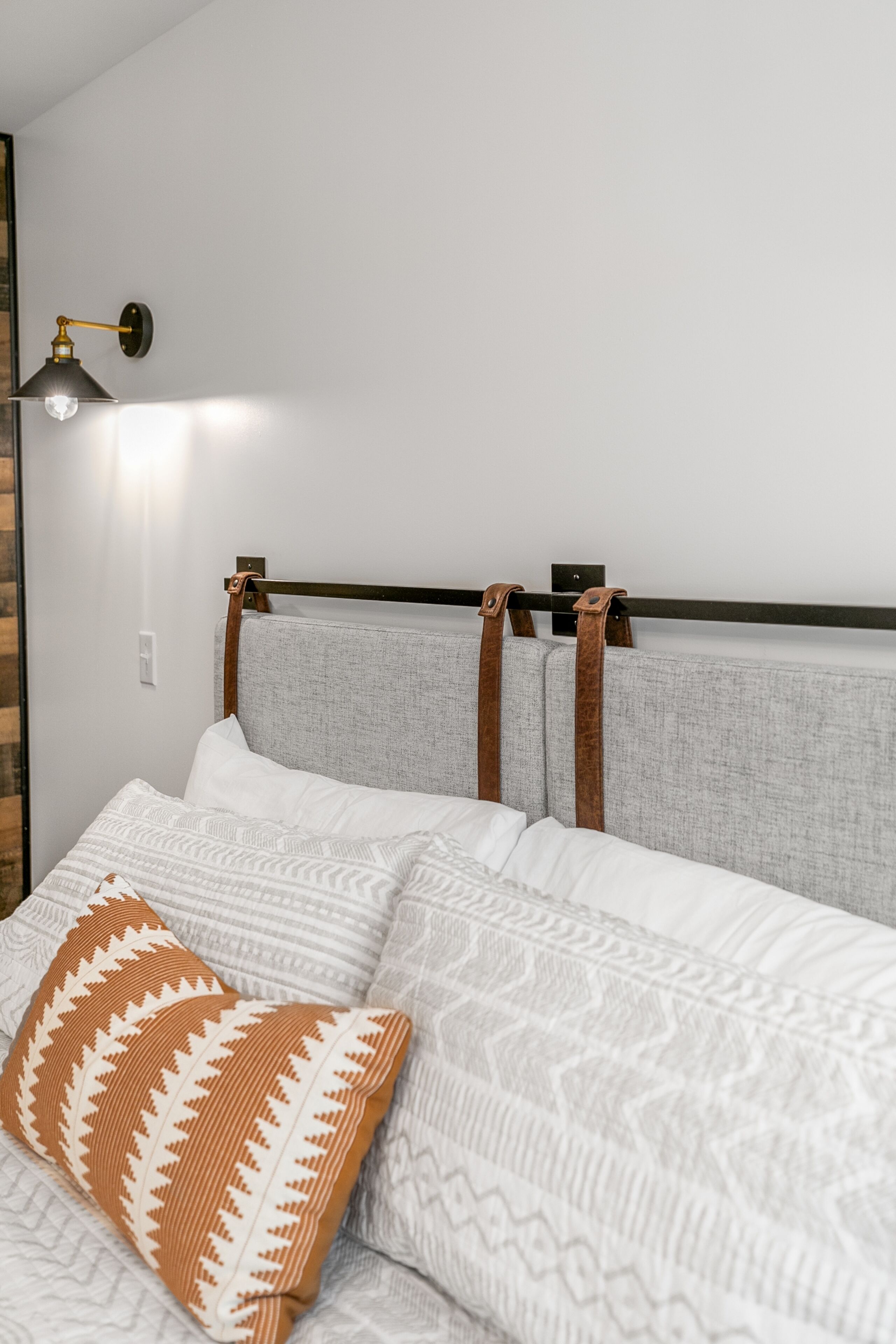 Una cama bien hecha con cabecera gris y tiras marrones, almohadas blancas y estampadas, y una lámpara fijada a la pared.