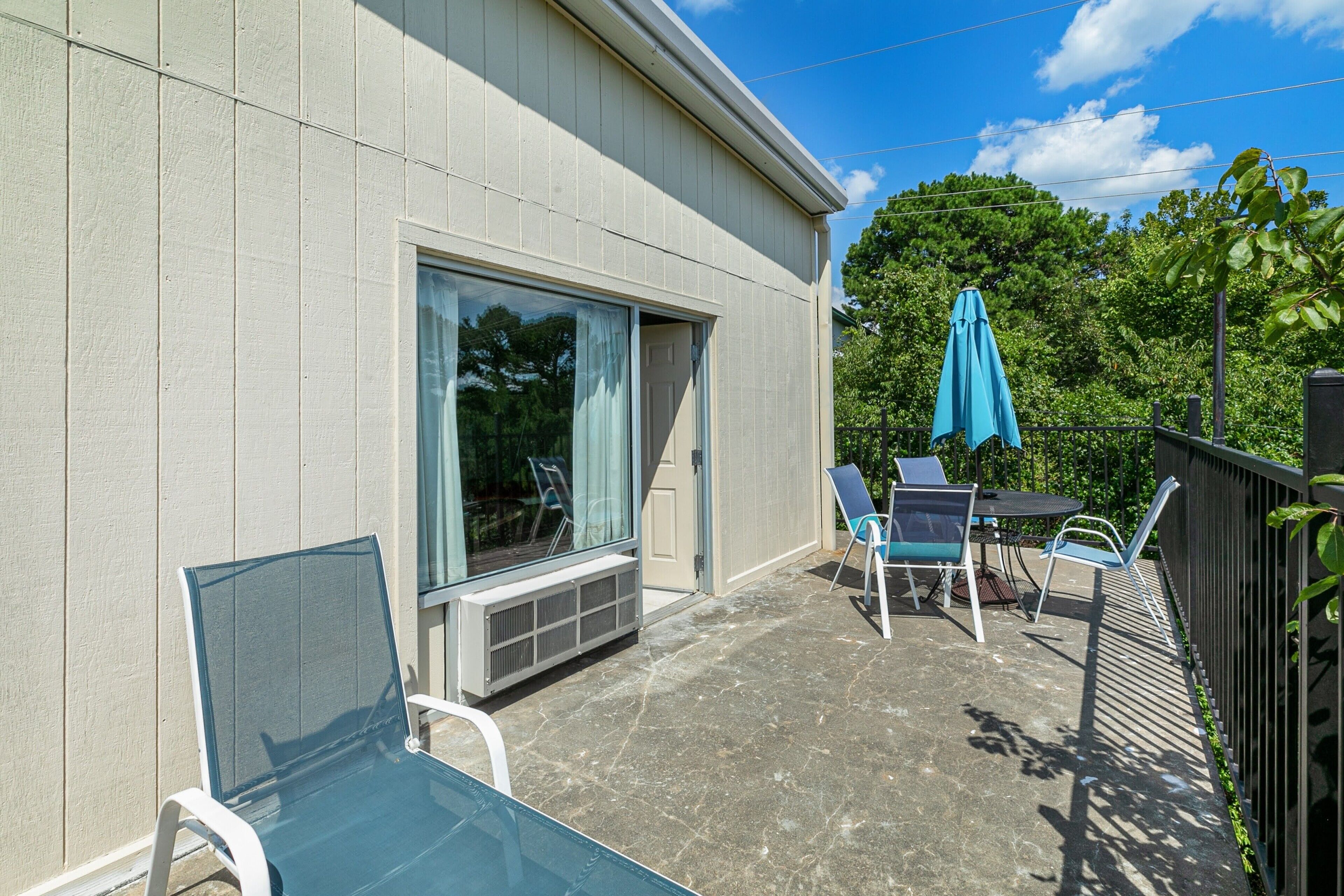 A patio with a table and chairs, a blue umbrella, and a window overlooking greenery.