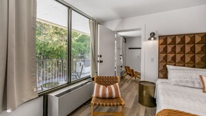Una habitación de hotel moderna con un ventanal grande, una silla de madera, una cama con ropa de cama estampada y un cabecero geométrico.