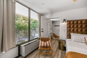 Una habitación de hotel moderna con un ventanal grande, una silla de madera, una cama con ropa de cama estampada y un cabecero geométrico.