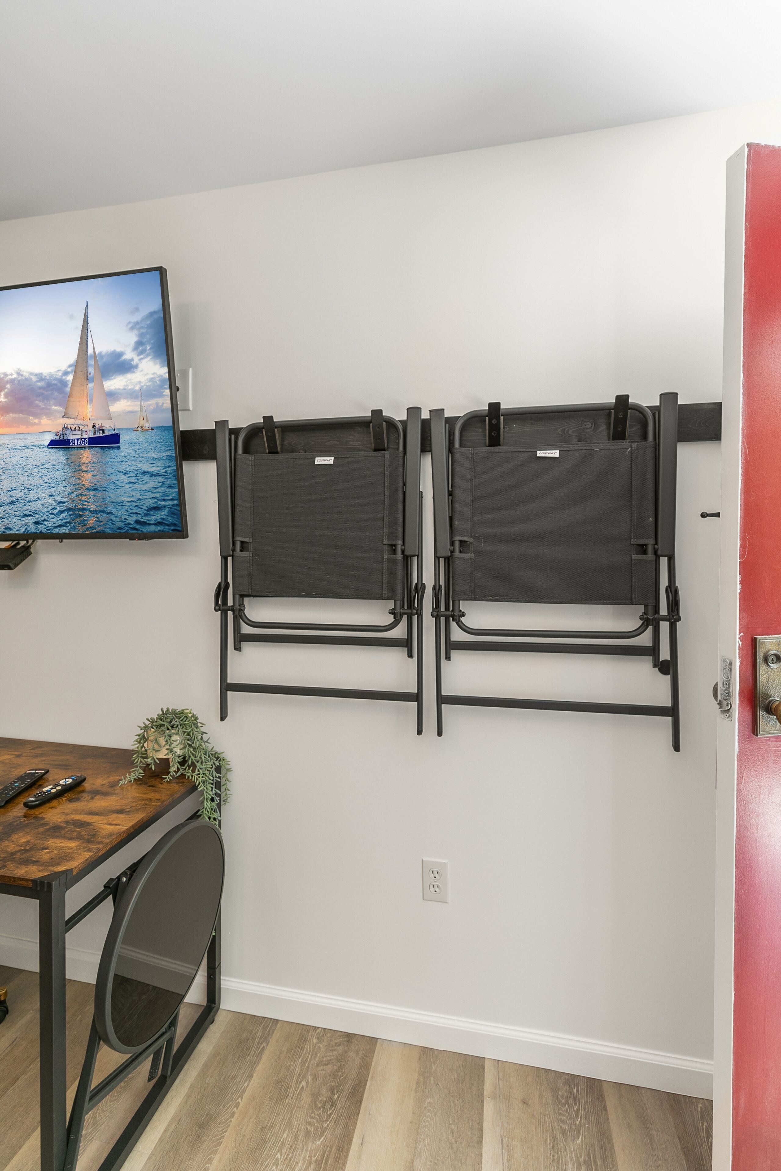A room with a desk, two wall-mounted chairs, a television displaying a sailboat, and a red door.