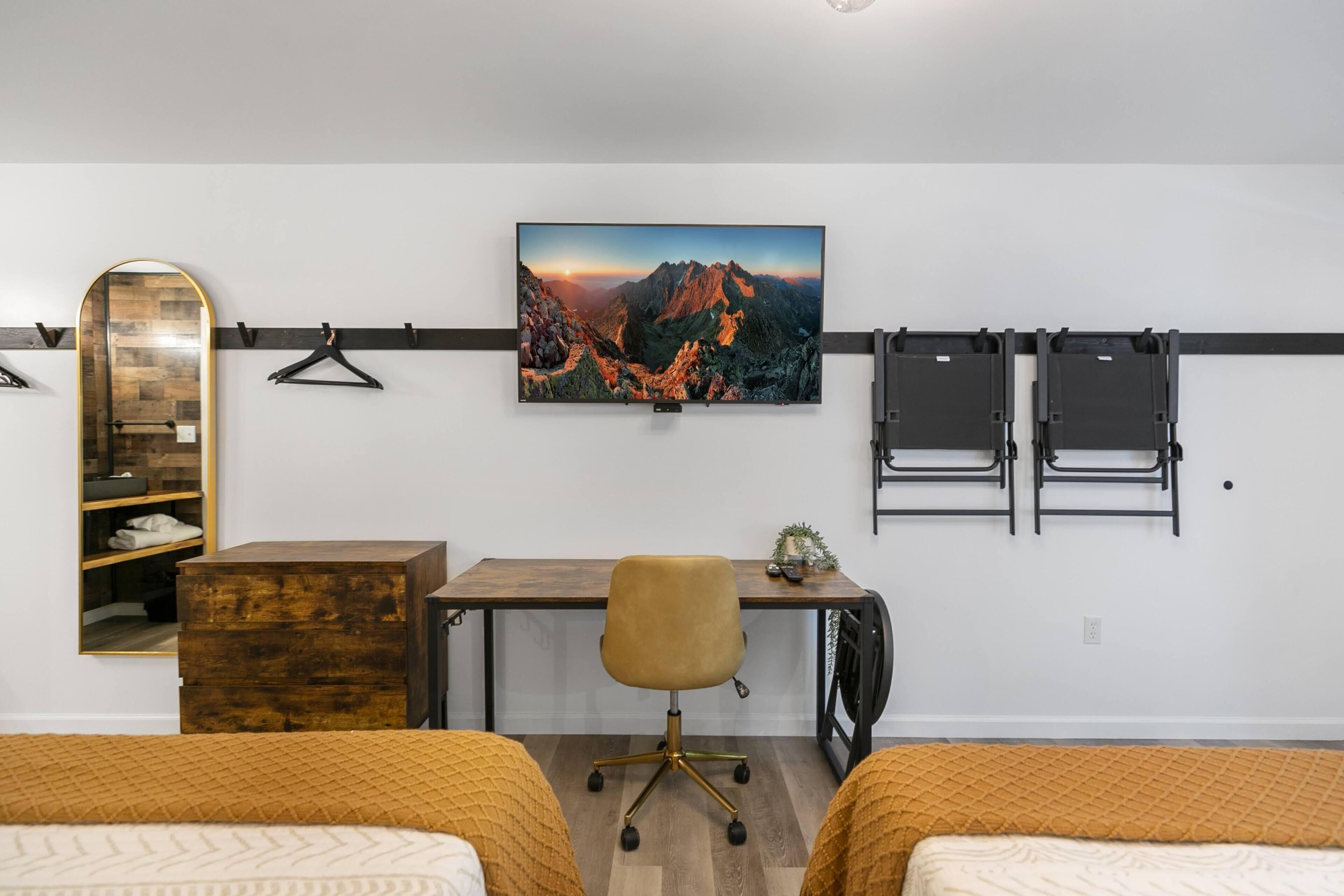 A bedroom with two beds, a desk, a chair, a mirror, and a wall-mounted TV displaying a mountain landscape.
