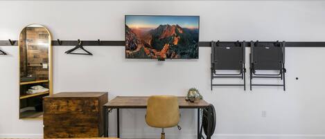 A bedroom with two beds, a desk, a chair, a mirror, and a wall-mounted TV displaying a mountain landscape.