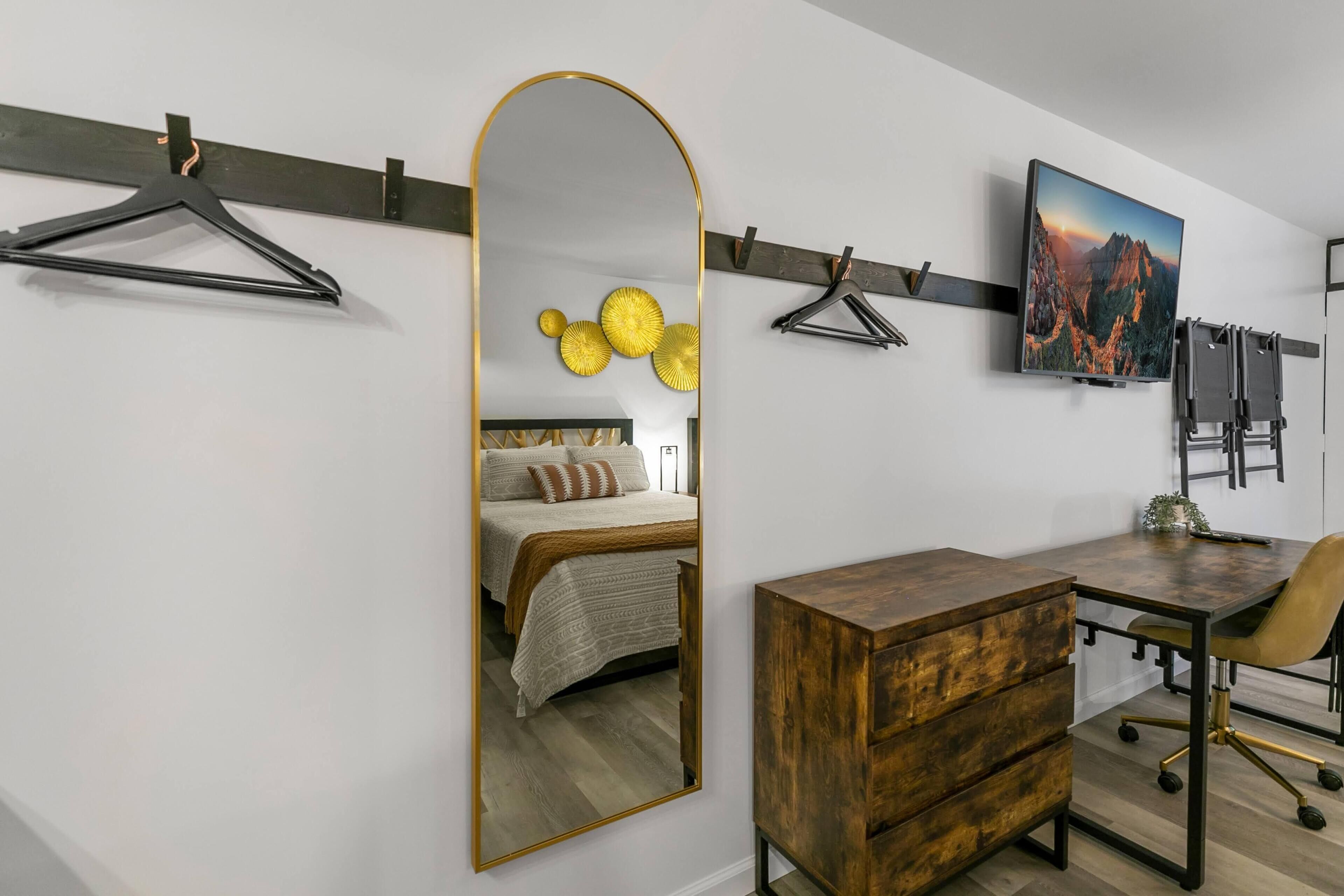 A bedroom with a bed, a wooden dresser, a desk with a chair, a large mirror, and a wall-mounted TV.