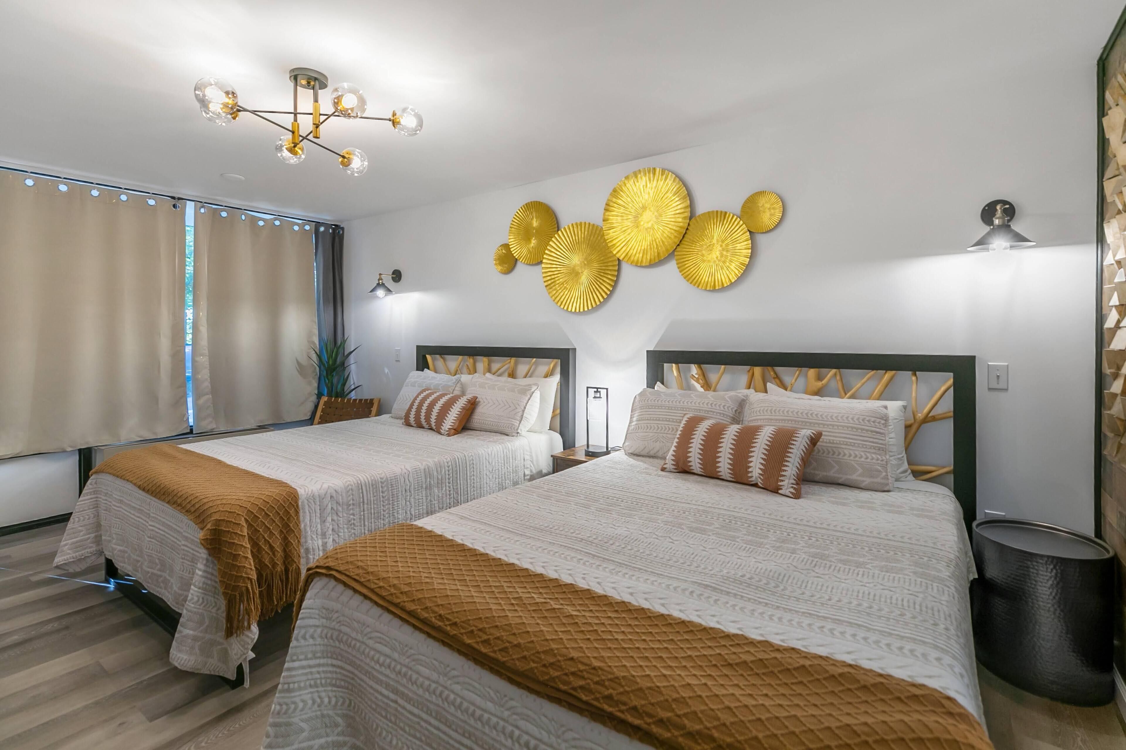 Quarto de hotel com duas camas, uma janela grande com cortinas, luminária fixada na parede e um objeto decorativo na parede.