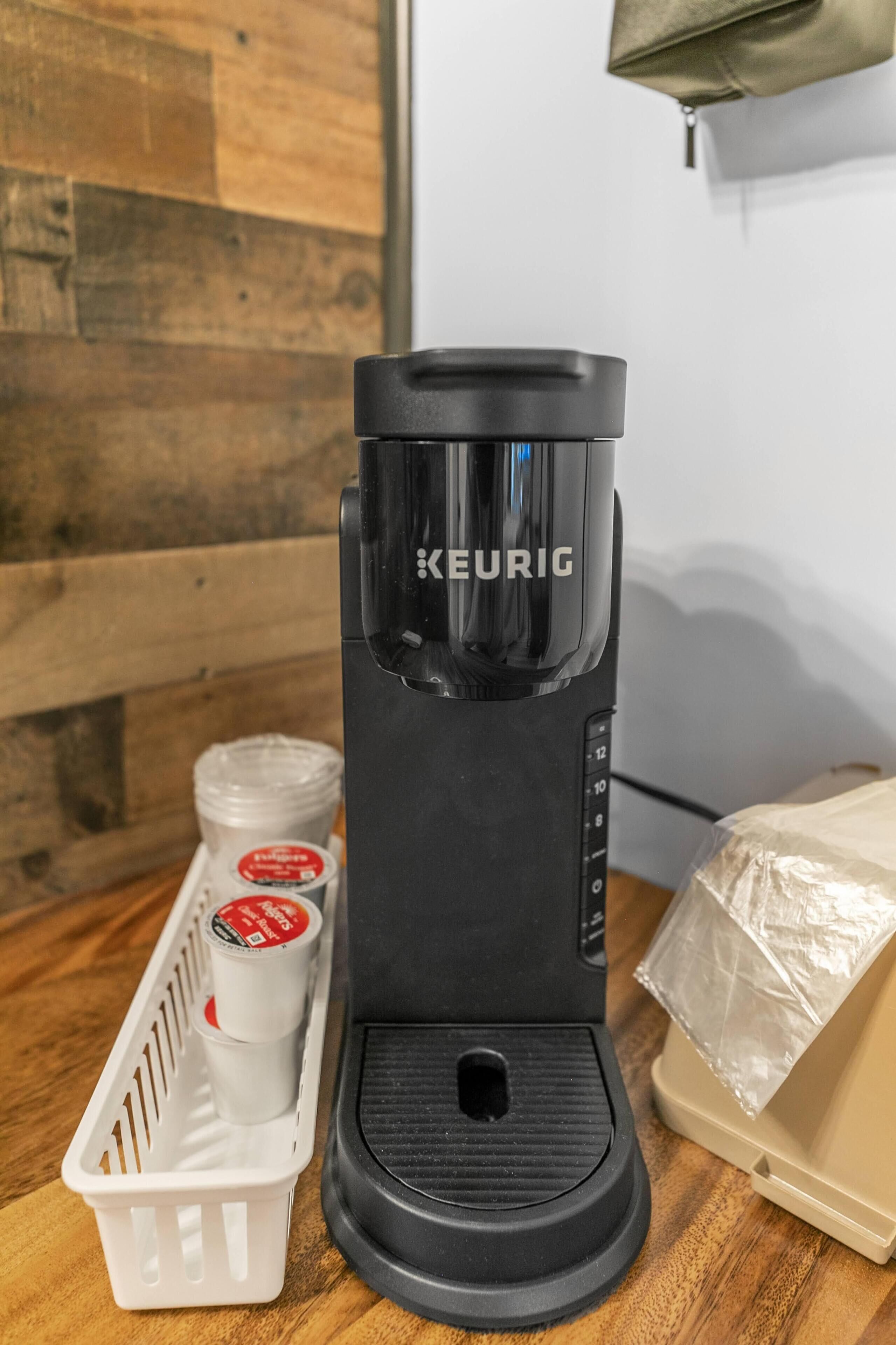 Une machine à café Keurig noire, posée sur une surface en bois, avec un panier blanc contenant deux tasses et un récipient.