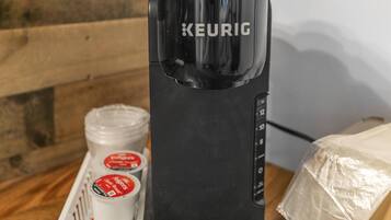 Une machine à café Keurig noire, posée sur une surface en bois, avec un panier blanc contenant deux tasses et un récipient.