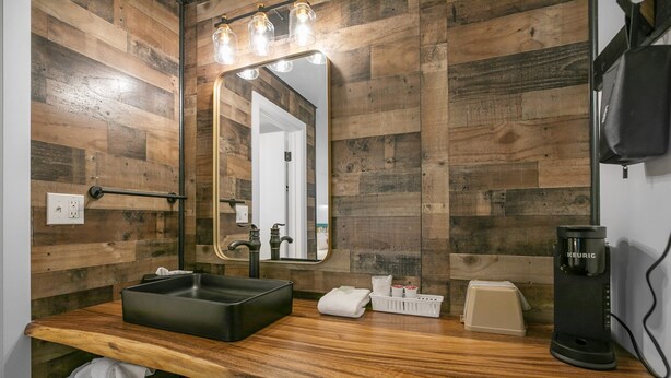 Un baño con pared de madera, un espejo grande, un lavamanos negro, una cafetera y un perchero para toallas.