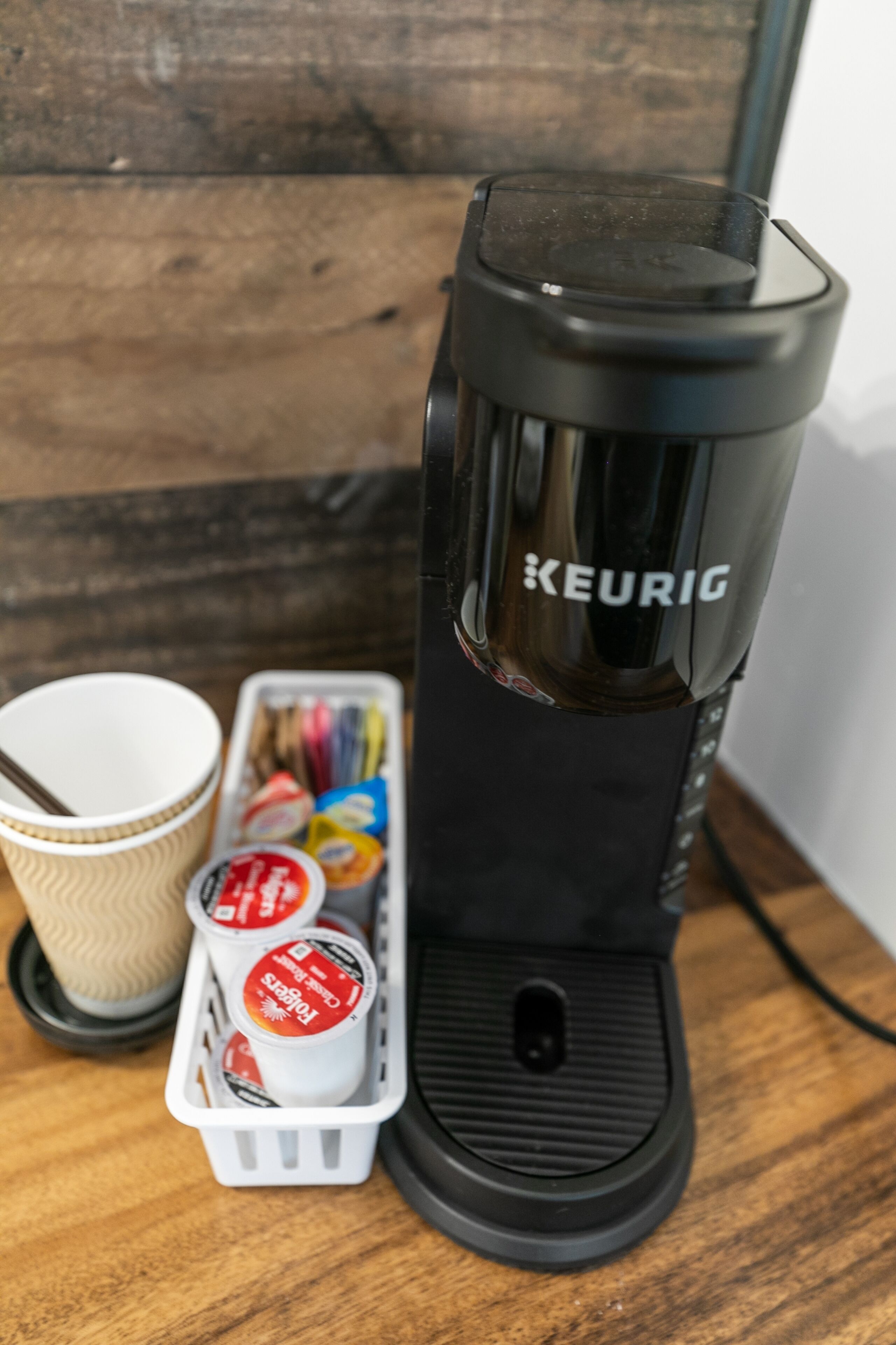 Eine Keurig-Kaffeemaschine auf einer Holztischplatte, zusammen mit einem Papierbecher und einer Kapselverpackung.