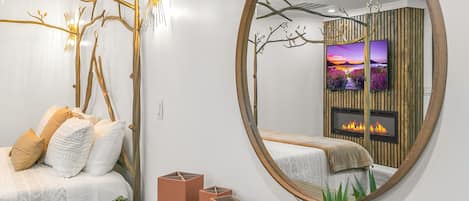 A modern bedroom with a large round mirror, a bed with white linens, a wooden headboard, and a bedside table with a clock and a plant.