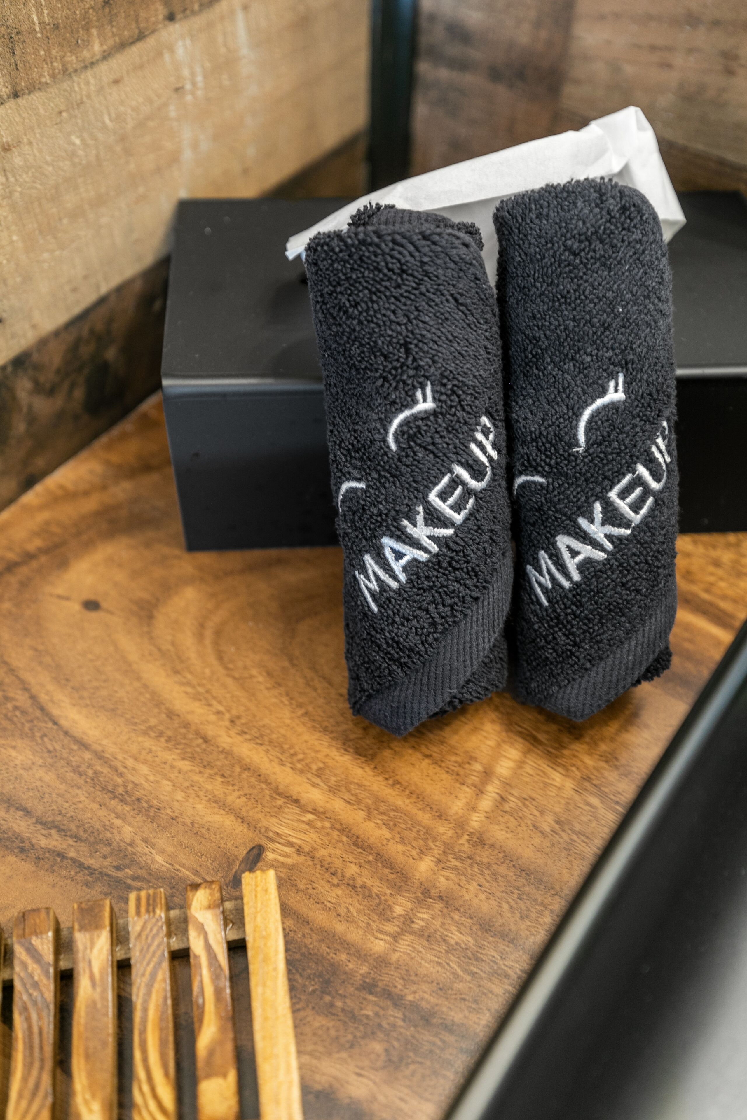 Duas toalhas pretas com a palavra 'MAKEUP' bordada, colocadas sobre uma superfície de madeira com uma caixa preta ao fundo.