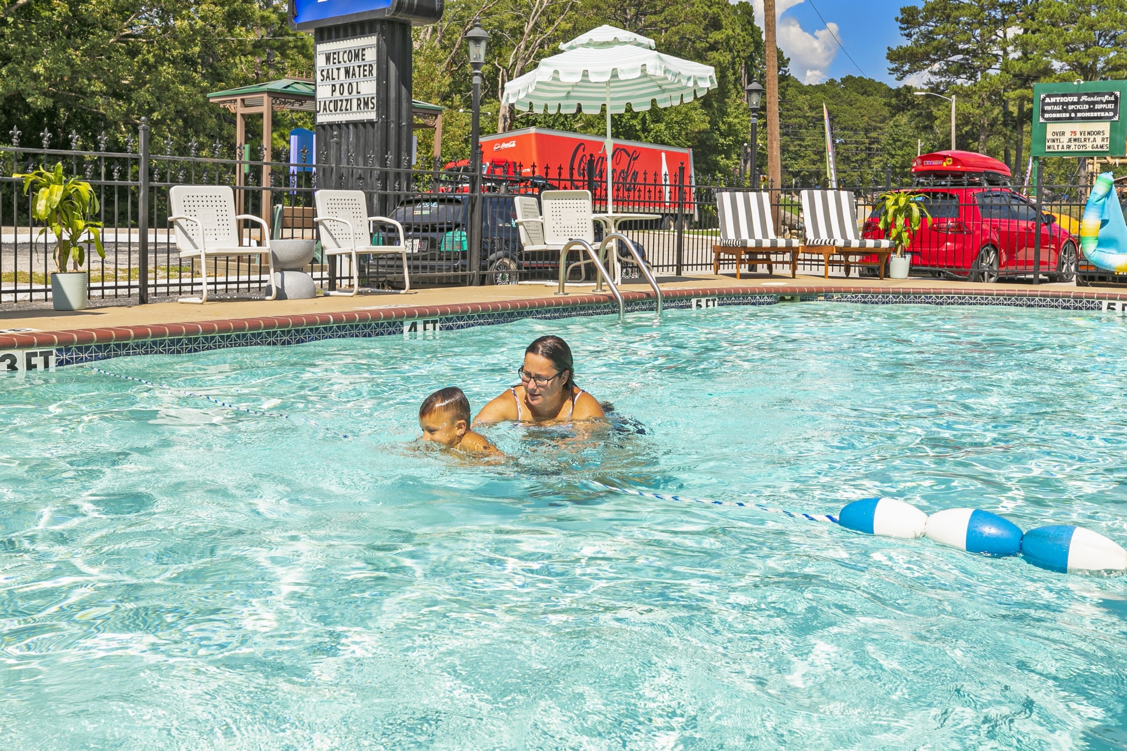 Uma piscina com uma fonte de água com a marca Coca-Cola, cadeiras de descanso e um caminhão Coca-Cola ao fundo.