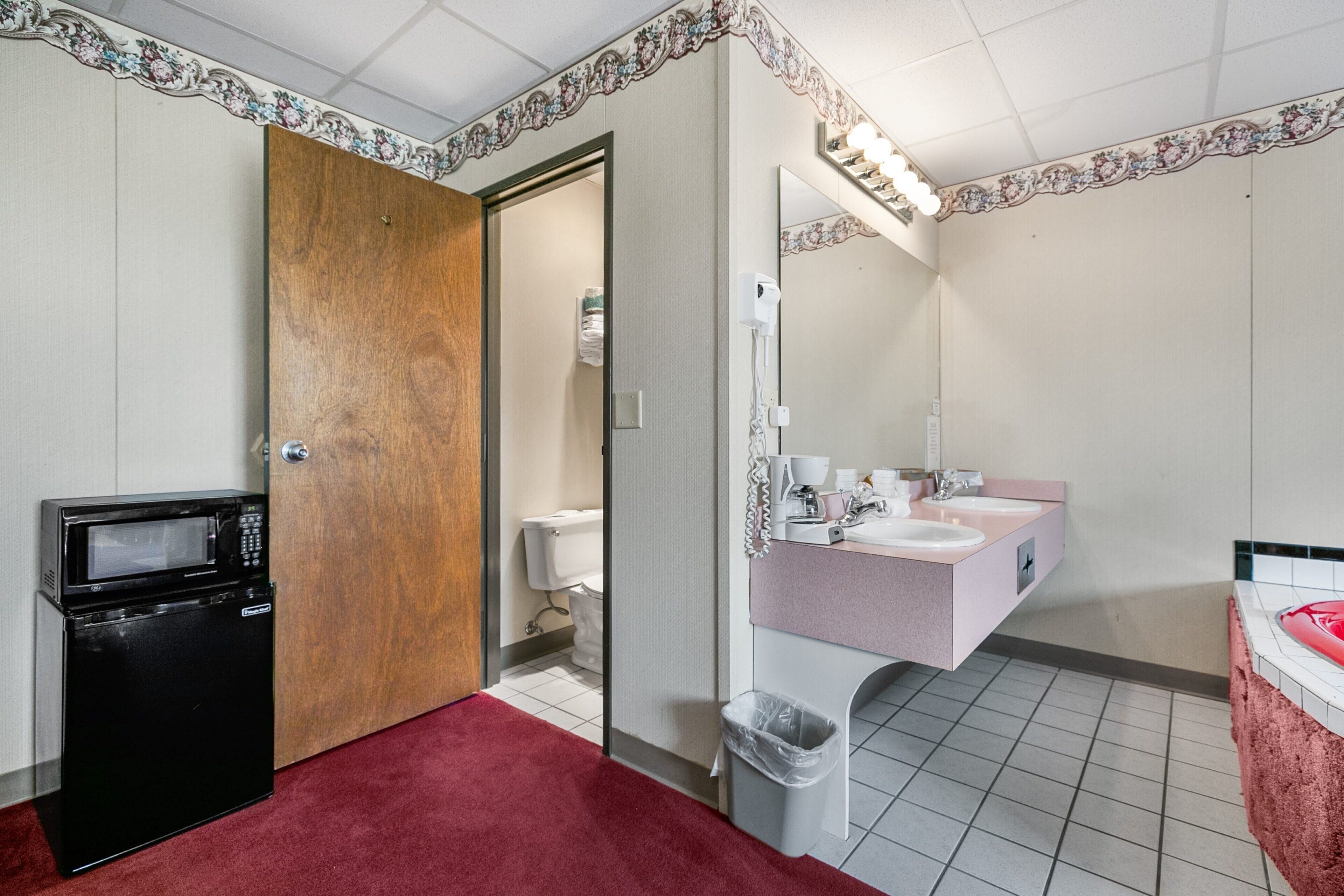 A compact hotel room with a microwave, a wooden door, a bathroom with a sink and mirror, and a red carpet.