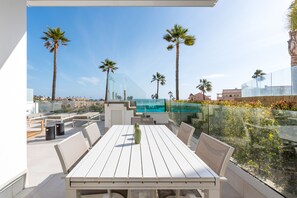 Terraço no telhado com mesa de jantar, cadeiras e guarda-corpo de vidro, com vista para uma piscina e palmeiras.