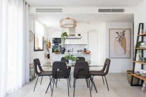 Uma área de refeições moderna com uma mesa de vidro e quatro cadeiras, uma luminária pendente e uma cozinha ao fundo.