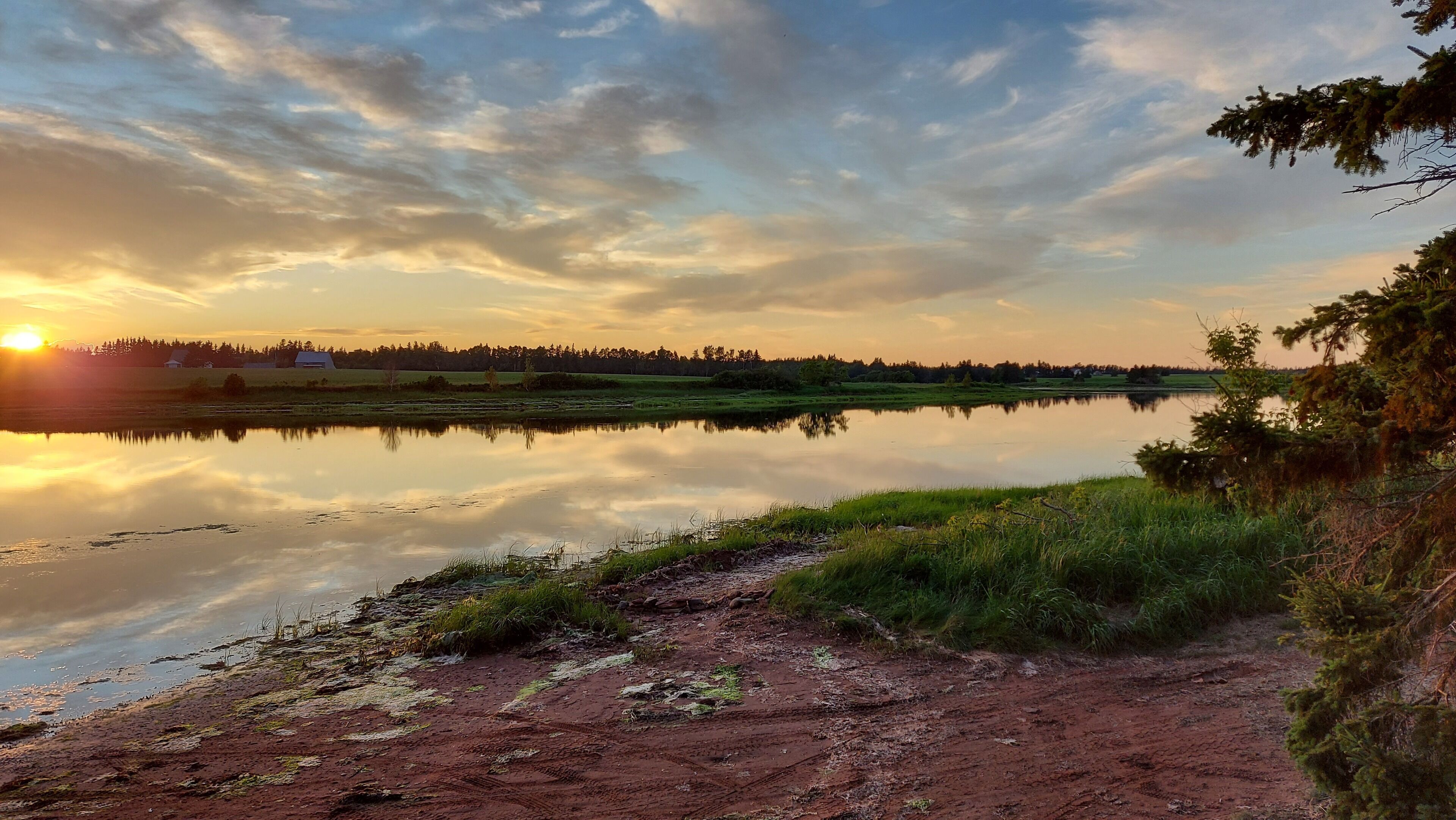 Vista serena de um lago ao pôr do sol, com águas calmas refletindo o céu e uma trilha de terra em direção à margem.