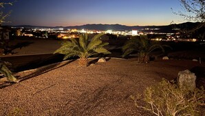 Vista noturna de uma cidade costeira, com um primeiro plano rochoso, palmeiras e luzes urbanas ao longe.