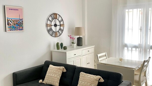 Un soggiorno moderno con un divano grigio scuro, un tavolo da pranzo bianco, una ventola a soffitto, una sveglia a parete e un quadro con una veduta urbana incorniciata.