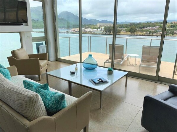 A modern living room with a glass table, beige sofas, and a view of a body of water and mountains. - Spectacular Oceanfront Penthouse, AC, Sleeps 6 (Lihue)