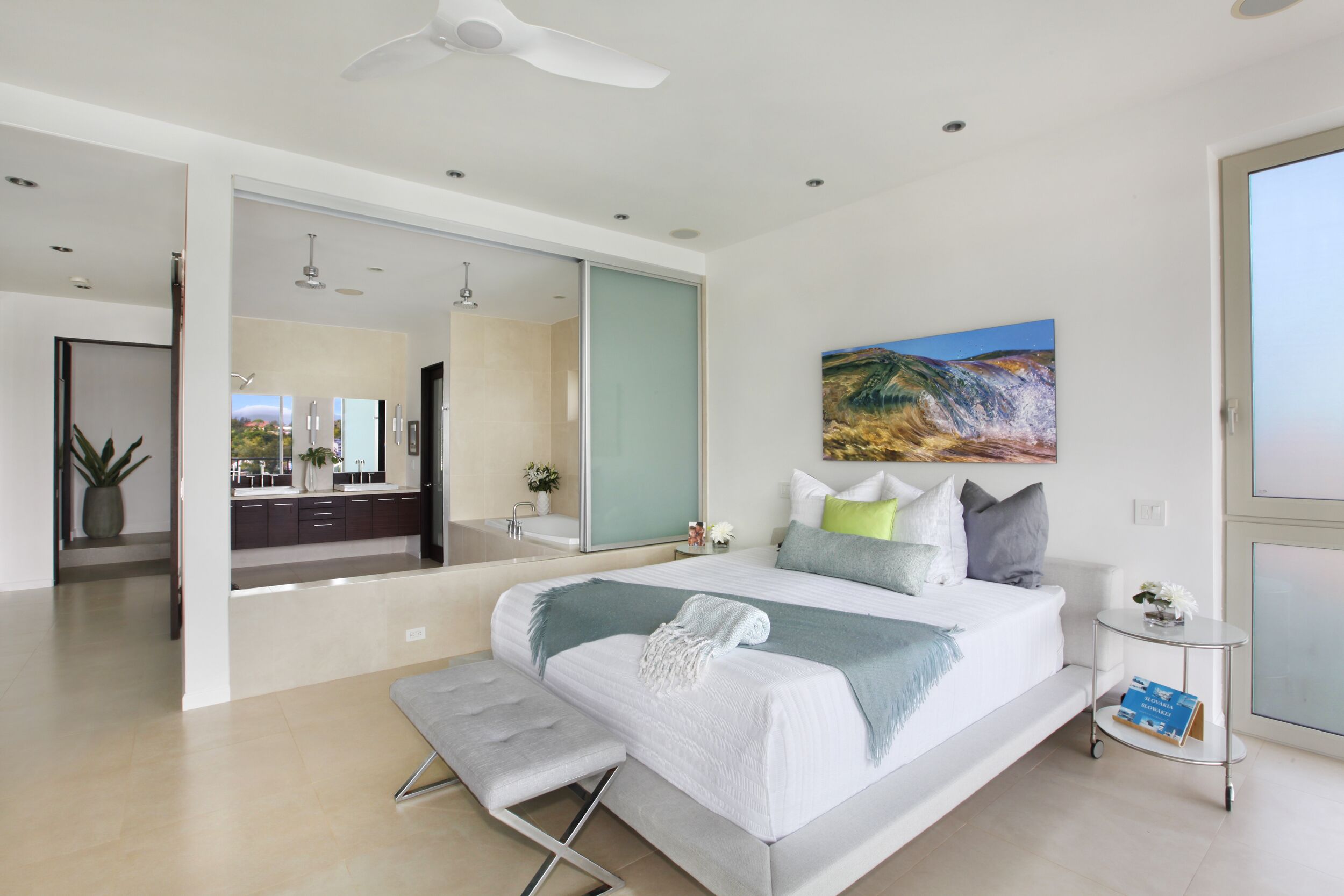 A modern bedroom with a large bed, a small bedside table, a ceiling fan, and a painting on the wall.