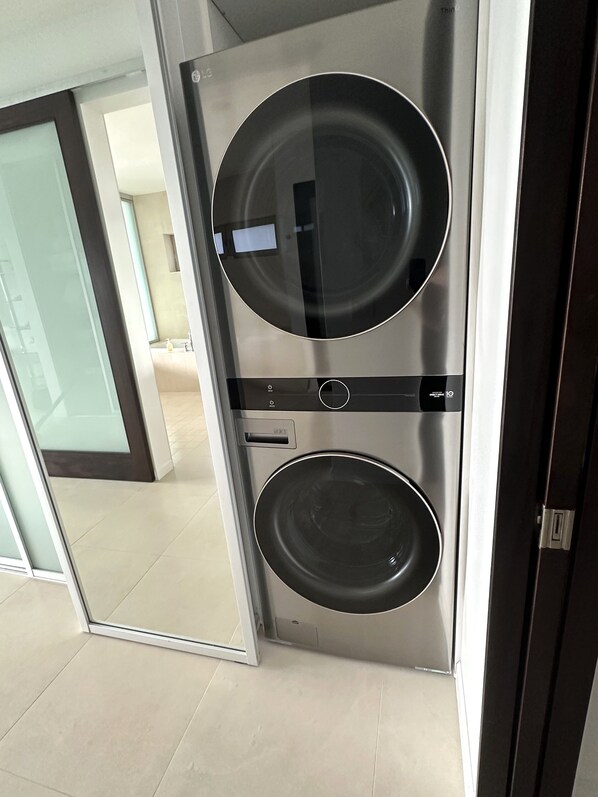 A compact, modern laundry room with a built-in washer and dryer, a control panel, and a glass door leading to a hallway. - Spectacular Oceanfront Penthouse, AC, Sleeps 6 (Lihue)