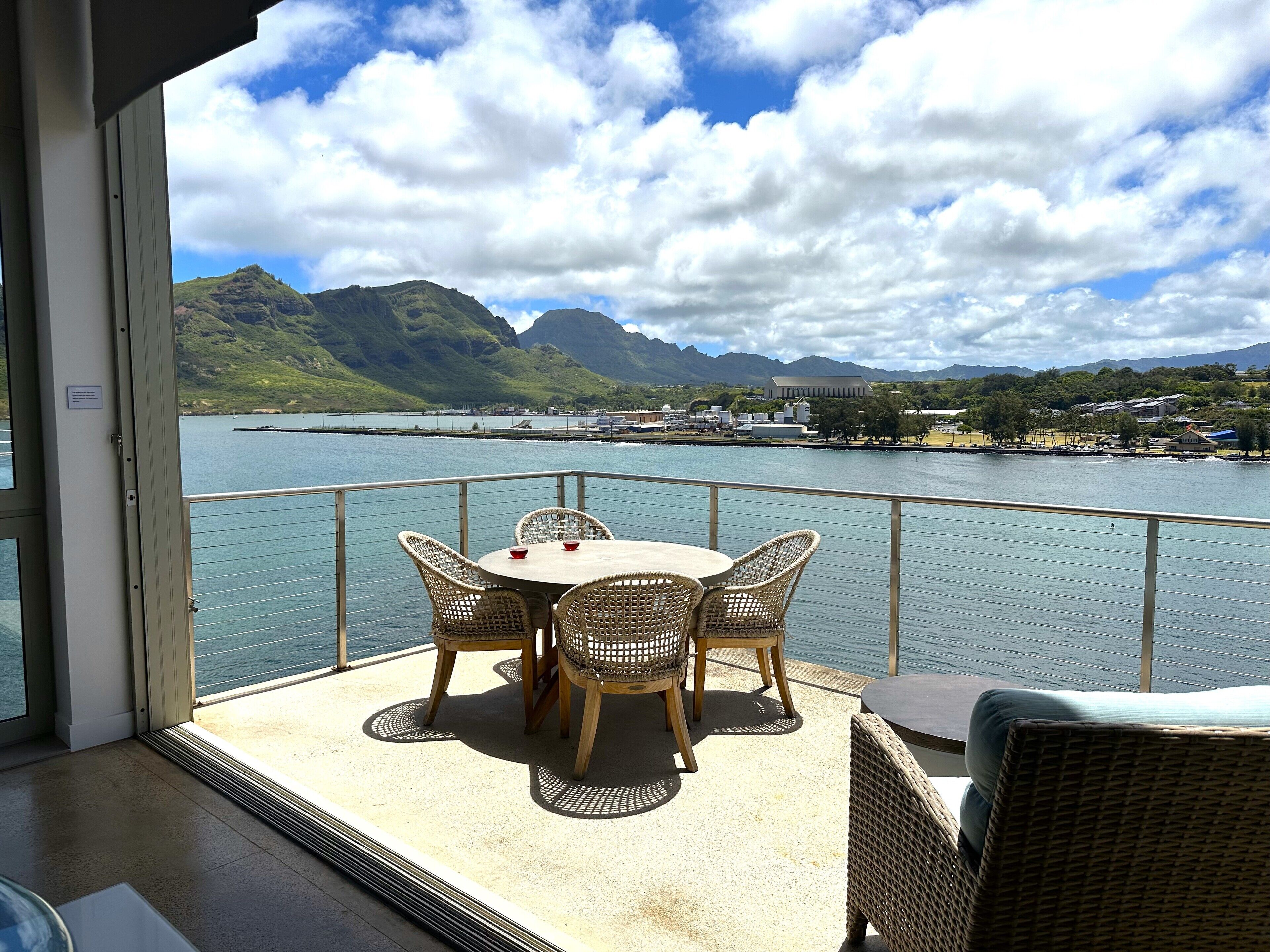 A balcony with wicker furniture overlooking a body of water, mountains, and a distant building.