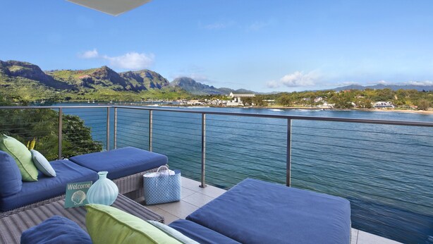 Un balcone con sedute imbottite blu, un vaso e un cartello di benvenuto, con vista su un lago e montagne.