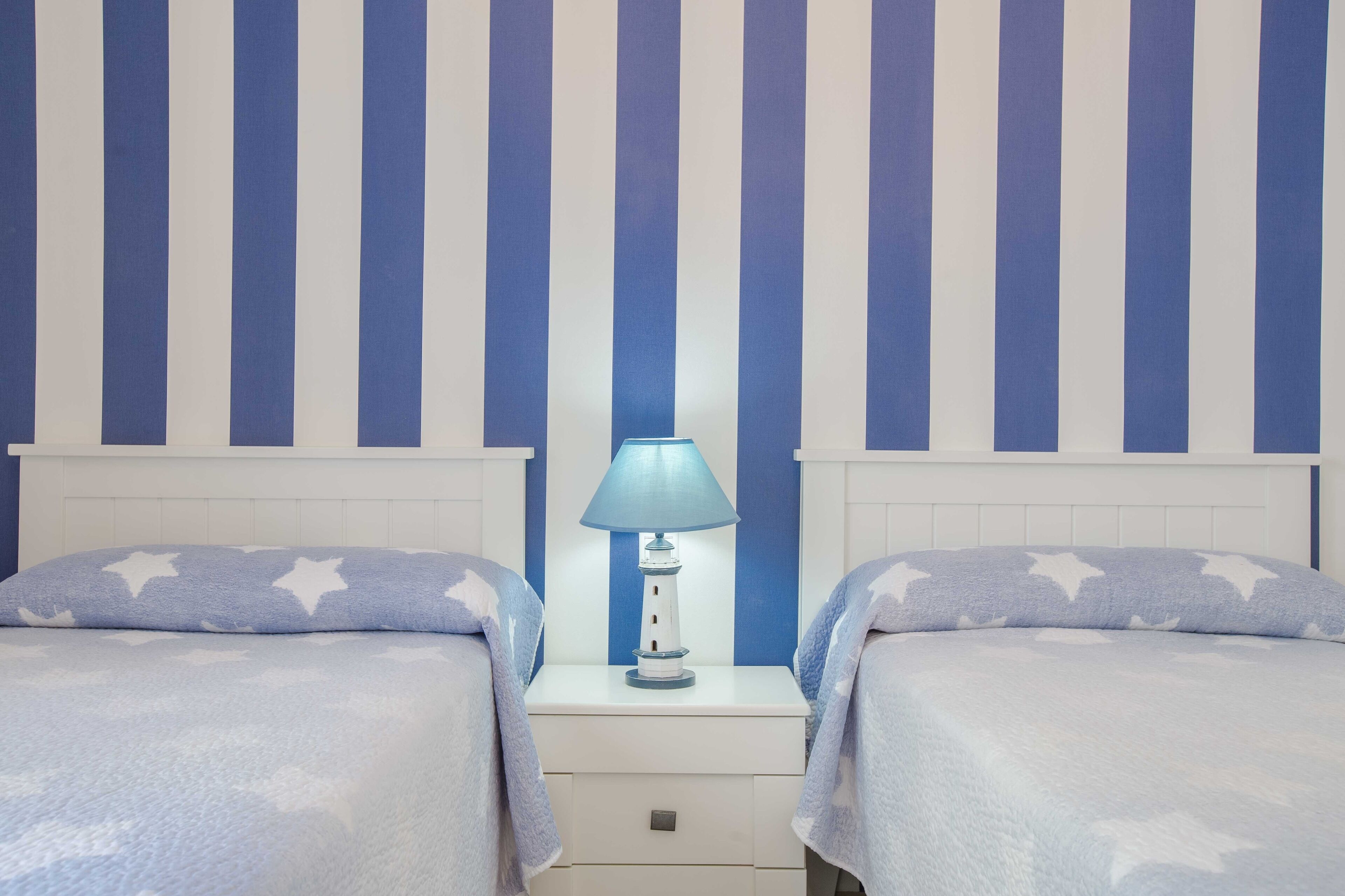 A bedroom with two beds, a nightstand with a lamp, and a striped wall.