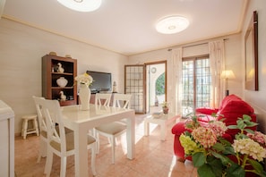 A dining area with a white table and chairs, a small side table with a vase of flowers, a red sofa, and a television in the background.