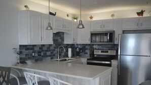 A modern kitchen with white cabinets, a central island with bar stools, stainless steel appliances, and decorative wall elements. - Beautiful views of beach, sunset, sunrise and the Blue Herons at the pond... (Pensacola)